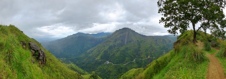 Panoramic view of green mountain ranges in Ella, Sri Lanka with a dirt hiking trail on the right, a lone tree, rocky outcrops on the left, and a winding road visible in the valley under cloudy skies