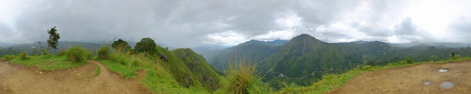 Panoramic mountain landscape showing green forested peaks and valleys in Ella, Sri Lanka under cloudy sky with dirt viewing area in foreground