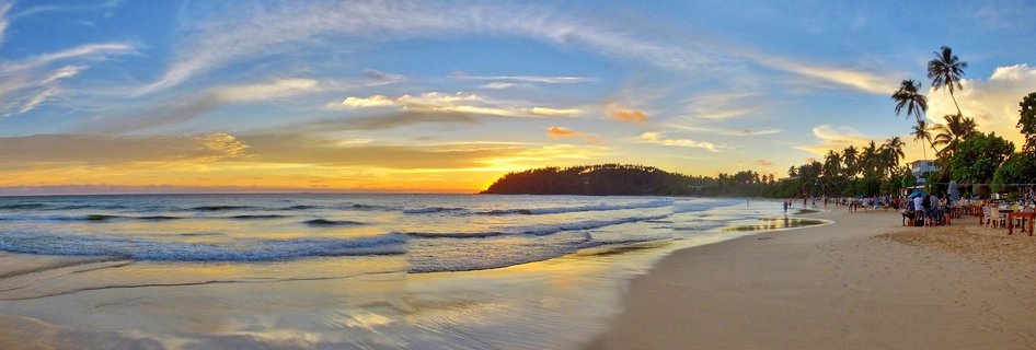 Panoramic beach sunset scene showing ocean waves, golden sky, palm trees, sandy shore with people at beachside tables, and a headland in the distance at Mirissa, Sri Lanka