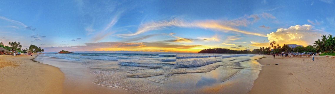 Wide beach scene at sunset showing ocean waves on wet sand, palm trees on both sides, people walking on the beach, and a colorful sky with orange and blue tones