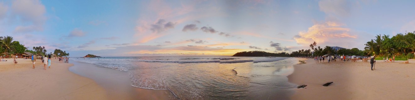 Panoramic beach scene at sunset showing ocean waves, golden sand, palm trees, scattered clouds with pink and orange colors, and people walking along Mirissa Beach in Sri Lanka