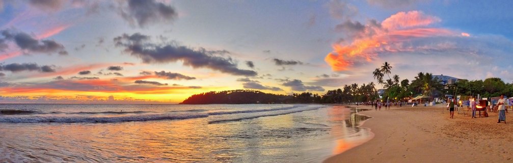 Panoramic beach sunset scene showing ocean waves, golden sand, palm trees, dramatic pink and orange clouds, and people walking along Mirissa Beach in Sri Lanka