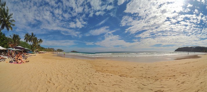 Panoramic beach scene showing golden sand, palm trees, beach umbrellas with people, turquoise ocean waves, blue sky with white clouds, and a distant headland at Mirissa, Sri Lanka