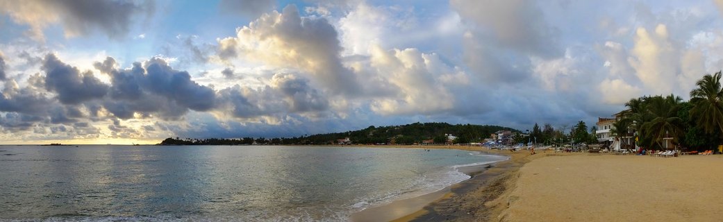 Wide panoramic beach scene showing Unawatuna Bay in Sri Lanka with ocean waves, sandy shore, palm trees, beachfront buildings, and dramatic cloudy sky at sunset