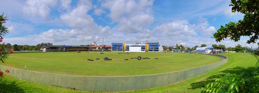 Wide sports field with green grass, black training tires scattered across it, concrete boundary wall, buildings in background, blue sky with white clouds, tropical trees framing the edges