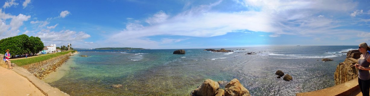Panoramic view of Galle Fort's stone coastal walls with turquoise ocean water, rock formations, colonial buildings, and visitors walking along the ramparts under a blue sky with scattered clouds