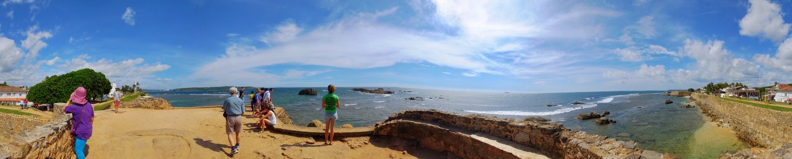 Panoramic view of tourists walking on stone fortification walls overlooking ocean with rocky coastline, blue sky with clouds, Galle Sri Lanka