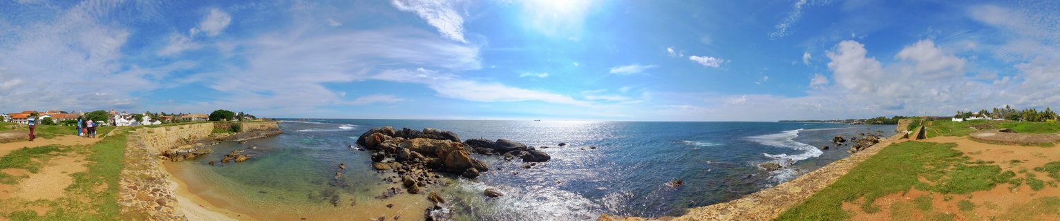 Panoramic view of historic Galle Fort stone walls extending along rocky coastline with blue Indian Ocean waters, waves, and clear sky with scattered clouds in Sri Lanka