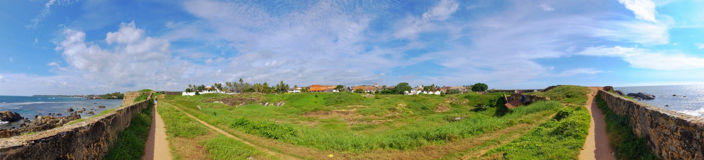 Panoramic photograph showing historic stone fort walls on both sides with green grassy field in center, ocean visible on left and right, blue sky with white clouds above, buildings and palm trees in distance