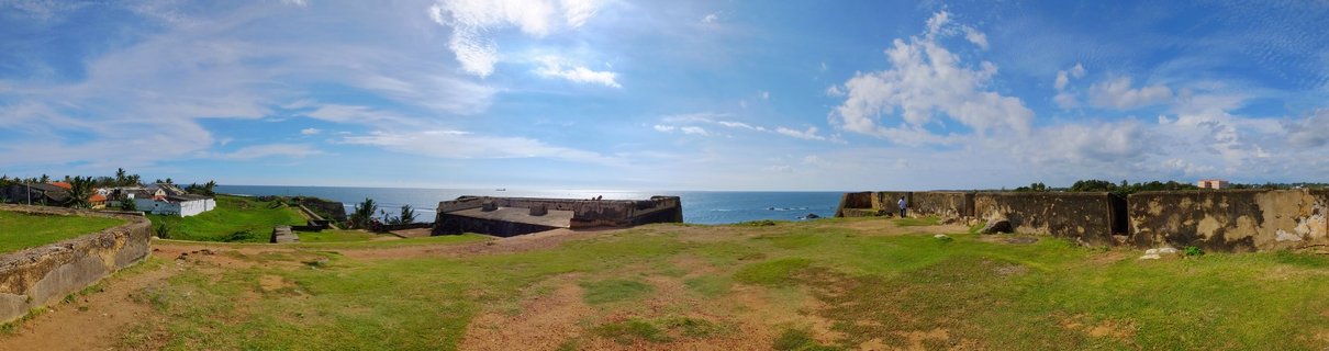 Panoramic view of historic stone fortification walls at Galle Fort with green grass foreground and Indian Ocean under blue sky with white clouds