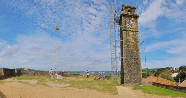 Stone clock tower with scaffolding and Sri Lankan flag on flagpole at Galle Fort with blue cloudy sky and harbor view in background