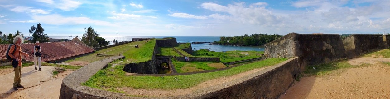 Panoramic view of historic Galle Fort showing grass-covered stone ramparts, colonial buildings with red-tiled roofs, two tourists walking on pathways, and the Indian Ocean coastline under a partly cloudy blue sky