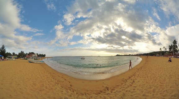 Wide panoramic view of Hikkaduwa Beach in Sri Lanka showing golden sand, turquoise ocean water, traditional fishing boats, palm trees, and people swimming during late afternoon with dramatic cloudy sky