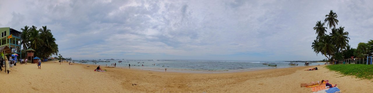 Wide beach panorama showing golden sand with people sunbathing and walking, palm trees on both sides, ocean with small boats, cloudy sky overhead, beachfront buildings visible on the left