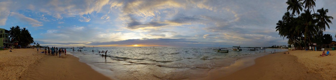 Wide beach panorama at sunset showing people on sand and in shallow water, fishing boats anchored offshore, palm trees on right, buildings on left, dramatic cloudy sky with setting sun on horizon