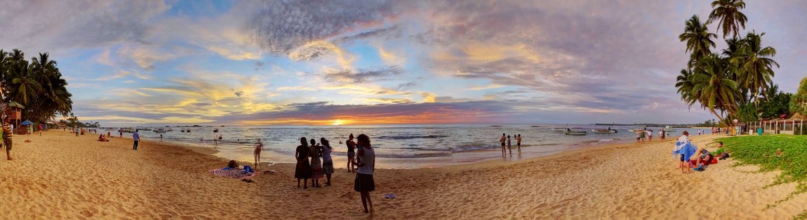 Wide panoramic beach scene at sunset showing people standing and sitting on golden sand, palm trees on both sides, fishing boats in the ocean water, and colorful clouds in orange pink and blue across the sky