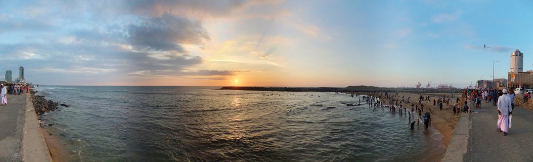 Panoramic view of sunset over the ocean at Galle Face Green beach in Colombo, Sri Lanka, with numerous people walking on promenades and wading in the water, city buildings and port cranes visible in the distance under a cloudy sky with golden sunlight