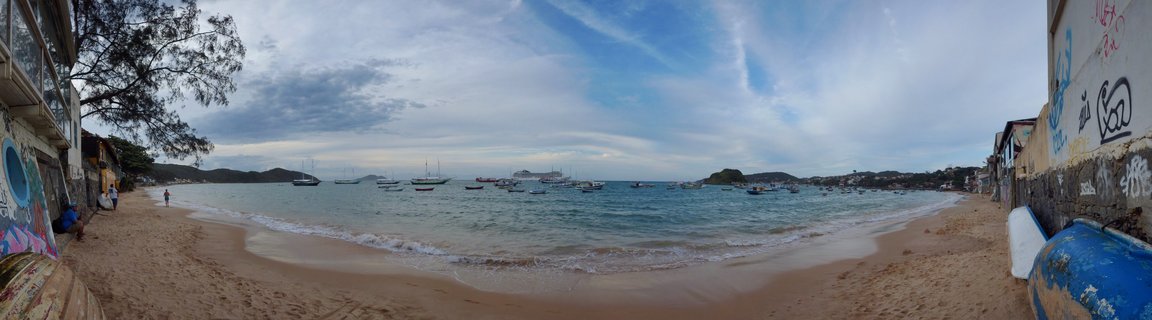Panoramic beach scene in Buzios Brazil showing a bay filled with anchored sailboats, sandy beach with gentle waves, buildings with colorful graffiti on both sides, and hills visible across the water under a partly cloudy sky