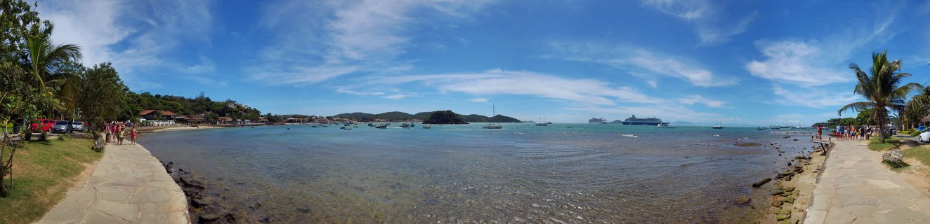 Panoramic beach scene showing a bay in Buzios Brazil with clear blue water, multiple boats and cruise ships, palm trees, stone walkways with people, and hills in the background under a partly cloudy sky