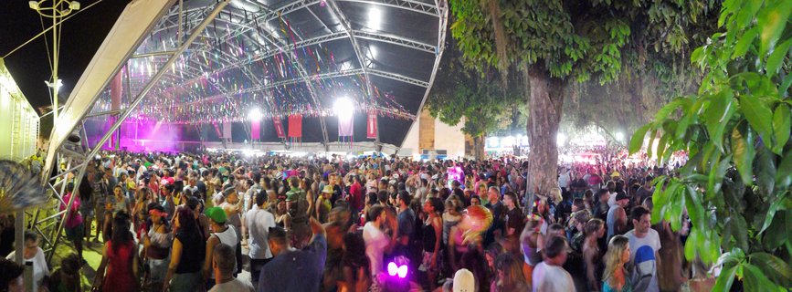 Large crowd of people at nighttime outdoor Carnival festival in Brazil, dancing under illuminated tent with colorful streamers and purple stage lighting