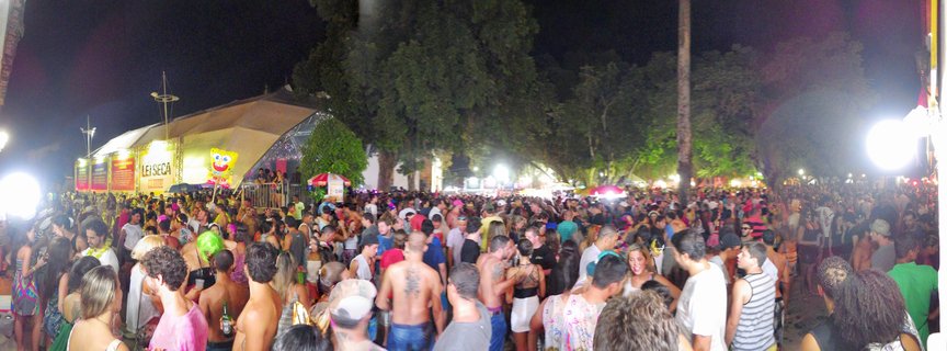 Large crowd of people at nighttime outdoor festival in Brazil with tent structure, stage lighting, and inflatable character visible