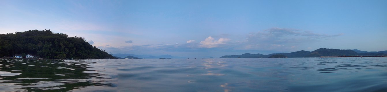Wide coastal panorama showing calm ocean water in the foreground with forested green hills on the left, distant mountains on the horizon, and blue sky with white clouds above Paraty Bay, Brazil