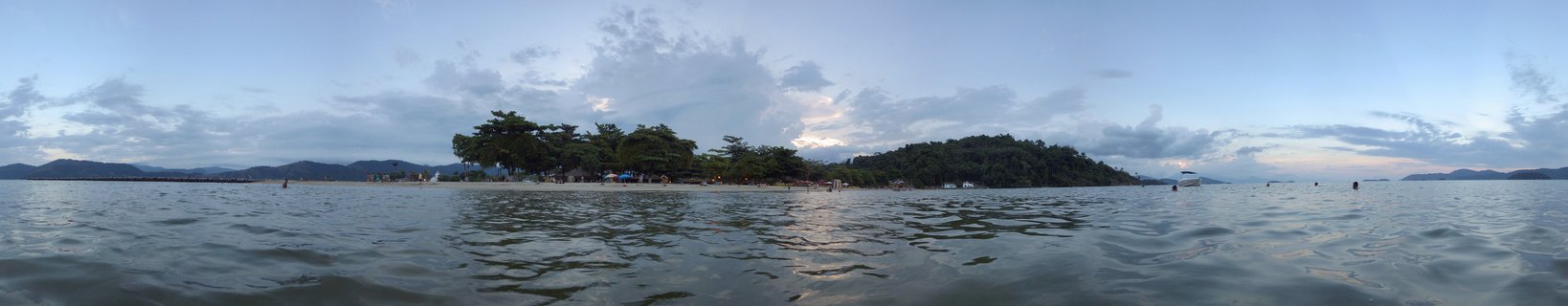 Panoramic coastal photograph showing calm bay waters in the foreground, a tree-covered island in the middle distance, and mountains along the horizon under a cloudy twilight sky