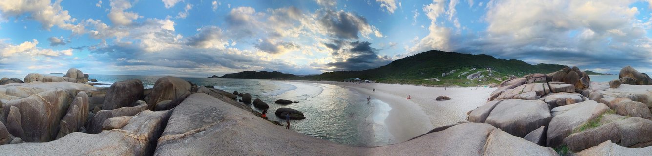 Panoramic view of large gray granite boulders in foreground framing a white sand beach with ocean waves, green hills in background, and dramatic cloudy blue sky at sunset in Florianopolis Brazil