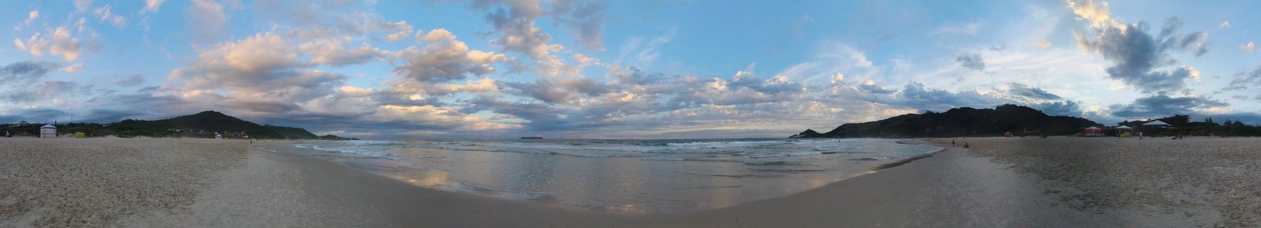 Wide panoramic beach scene at sunset in Florianopolis, Brazil showing wet sand with sky reflections, ocean waves, distant hills, and cloudy sky with golden hour lighting