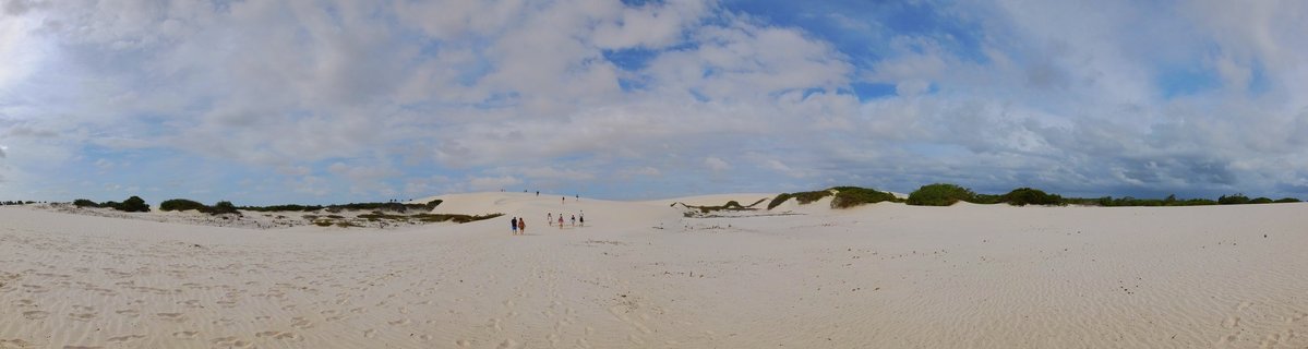 Panoramic view of white sand dunes with small groups of people walking across the landscape, green vegetation scattered throughout, under a cloudy blue sky in Barreirinhas, Brazil