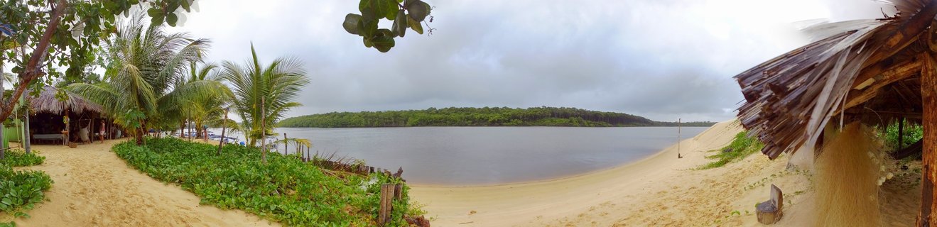 Panoramic view of a Brazilian river mouth with sandy beach, thatched-roof huts, palm trees, tropical vegetation, and calm water under cloudy sky
