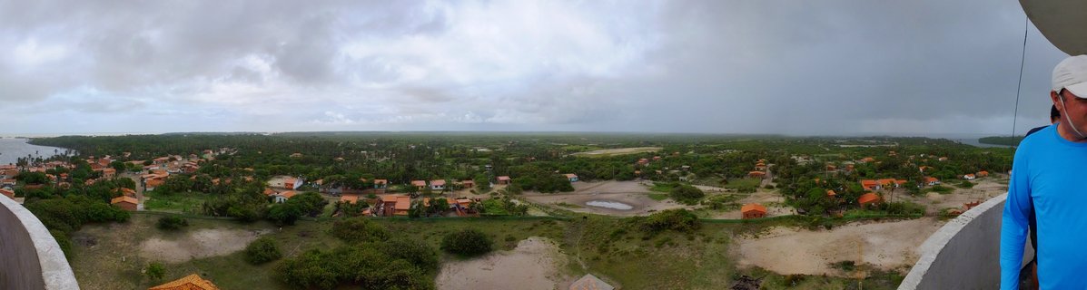 Wide aerial panorama showing a Brazilian coastal village with orange-roofed houses scattered among green tropical vegetation under cloudy skies
