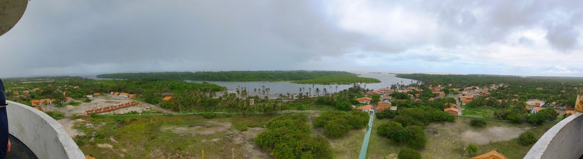 Panoramic aerial view showing a tropical coastal village in Brazil with orange-roofed houses, palm trees, dense green vegetation, and a winding river under cloudy skies, photographed from an elevated concrete tower structure