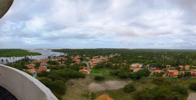 High-angle view of a Brazilian coastal village showing orange-roofed houses among green tropical trees, with a river flowing toward the ocean under cloudy skies, white curved structure visible in foreground.