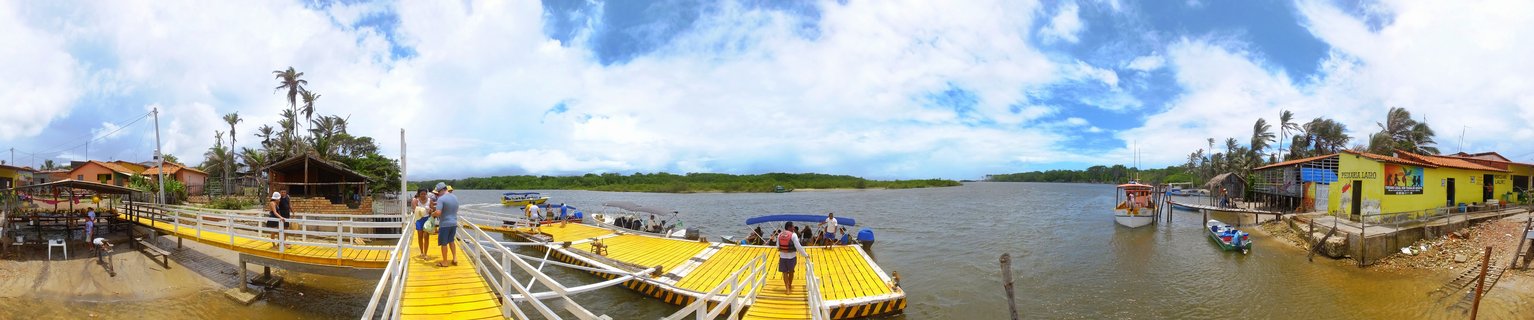 Panoramic waterfront scene showing a yellow wooden pier with people walking, small boats moored in calm water, colorful buildings along the shore, and palm trees under a blue sky with white clouds