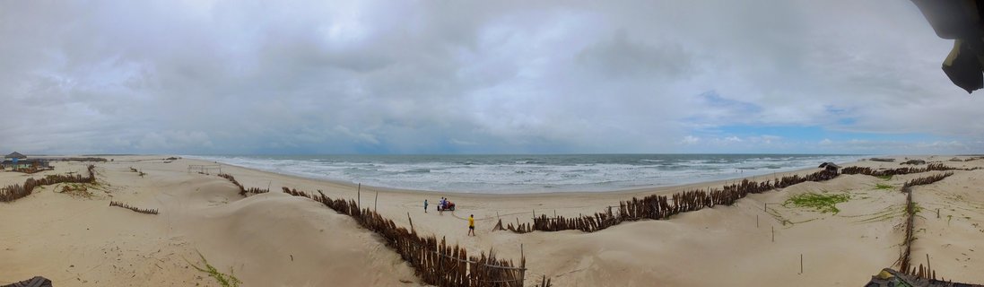 Wide panoramic beach scene showing sand dunes with wooden erosion control fences, ocean waves, and overcast sky in São Luís, Brazil with a few people visible on the beach
