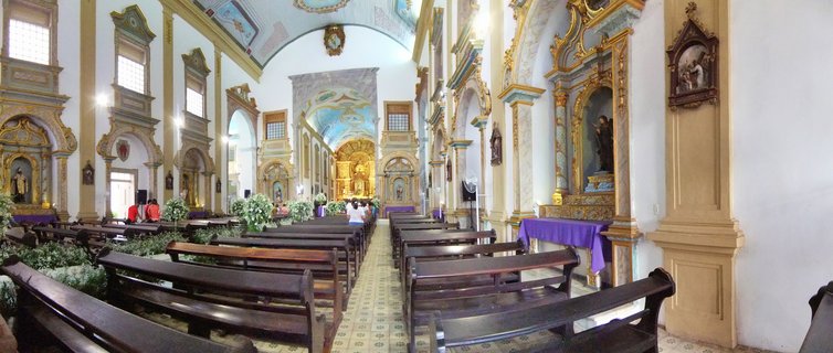 Panoramic interior of a Brazilian colonial church showing rows of dark wooden pews, white walls with gold ornate altars, arched doorways, painted ceiling, and purple altar cloths with people visible in the background