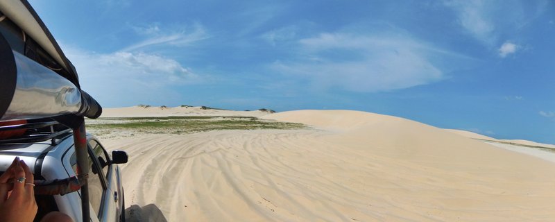 Silver four-wheel-drive vehicle on white sand dunes with passenger hands visible on roll bar, blue sky with clouds above, sparse green vegetation on distant dune ridges in Jericoacoara Brazil