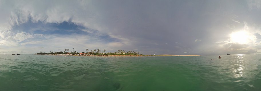 Panoramic coastal photograph showing turquoise ocean water in the foreground, a strip of land with palm trees and buildings in the middle distance, and a dramatic cloudy sky with sun visible on the right