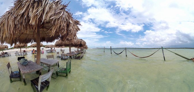 Beach restaurant with thatched roof umbrellas, wooden tables and chairs in shallow turquoise water, hammocks suspended over lagoon, blue sky with clouds, Jericoacoara Brazil