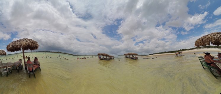Panoramic view of tourists sitting in chairs and hammocks in shallow tropical lagoon water with multiple thatched-roof beach structures under cloudy blue sky in Jericoacoara, Brazil