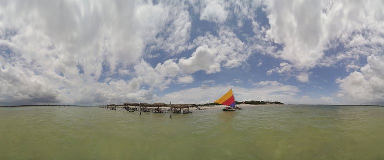 Panoramic view of a colorful sailboat on shallow green water with wooden pier structures and dramatic cloudy sky in Jericoacoara, Brazil