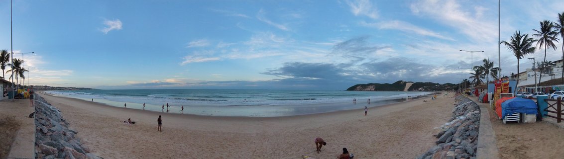 Wide panoramic beach photograph showing sandy shore with people, palm trees on both sides, ocean waves, blue sky with clouds, and coastal dunes in the distance at Natal, Brazil