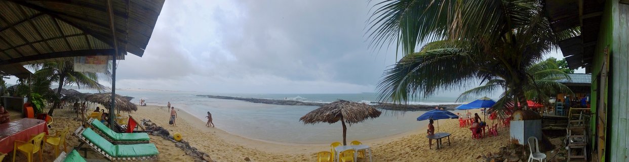 Panoramic view of Praia da Pipa beach in Brazil showing sandy shoreline with thatched umbrellas, blue beach umbrellas, yellow chairs, palm trees, beach structures with thatched roofs, people walking on sand, and ocean with waves under cloudy sky