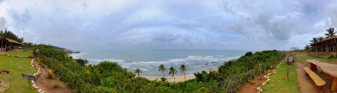 Wide panoramic view of Praia da Pipa beach in Brazil showing ocean waves, sandy shore, palm trees, green tropical vegetation, and wooden beachfront buildings under cloudy sky