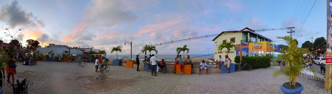 Wide panoramic view of a paved beachfront plaza in Praia da Pipa, Brazil at dusk with people sitting on colorful benches, palm trees in planters, buildings with shops, and the ocean visible in the background under a cloudy sky with pink sunset colors