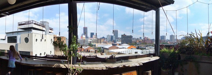 View from a rooftop bar showing Johannesburg city skyline with buildings, hanging light bulbs, wooden bar counter, plants, and blue sky with clouds