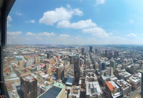 High-angle panoramic view of Johannesburg city center showing numerous skyscrapers and urban buildings under a blue sky with white clouds, viewed from an observation deck