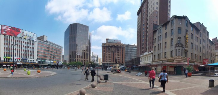 Wide urban plaza in Johannesburg city center with pedestrians walking, surrounded by modern and historic buildings under blue sky with clouds