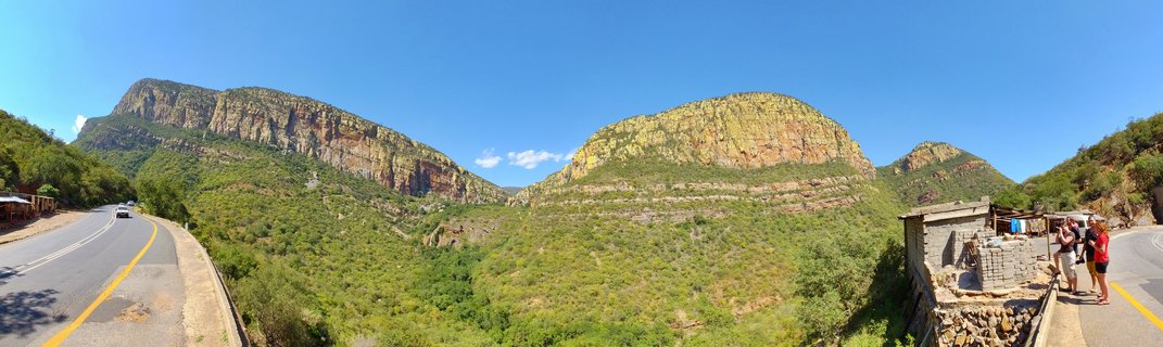 Panoramic landscape showing steep rocky cliffs covered with green vegetation, a winding road with a white vehicle, and tourists at a viewpoint under clear blue sky
